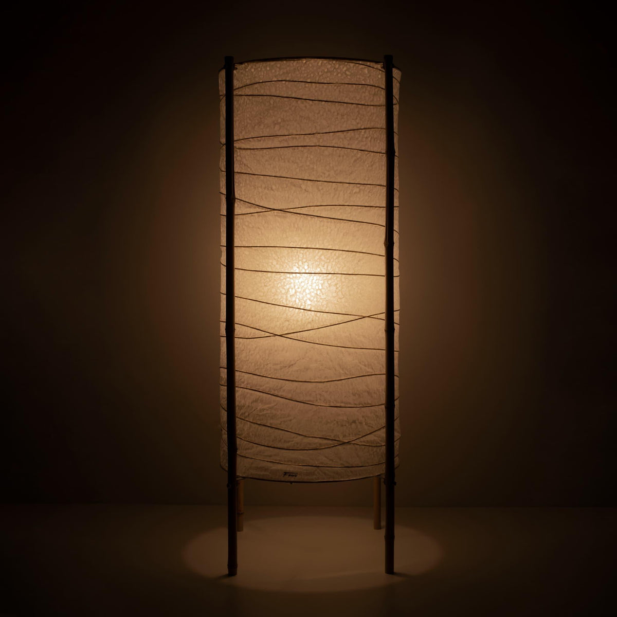 Full view of a cylindrical Japanese lamp with washi paper shade and bamboo frame, warm glow on brown background