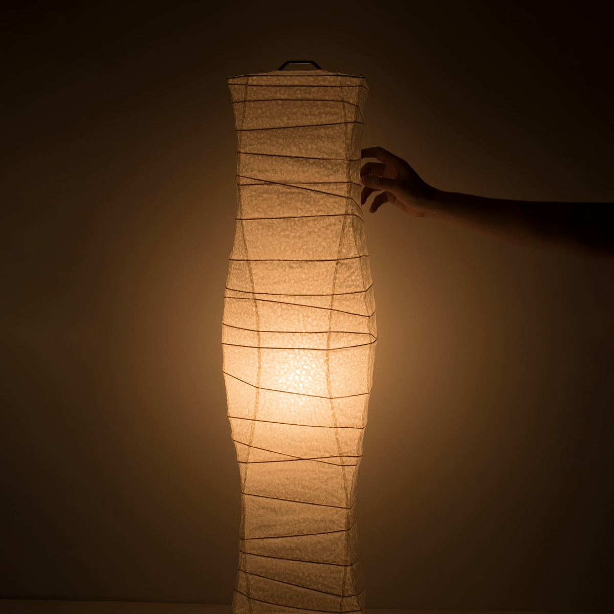 "Une main touche délicatement le côté d'une lampe en papier japonais washi aux courbes fluides, émettant une douce lumière chaude"