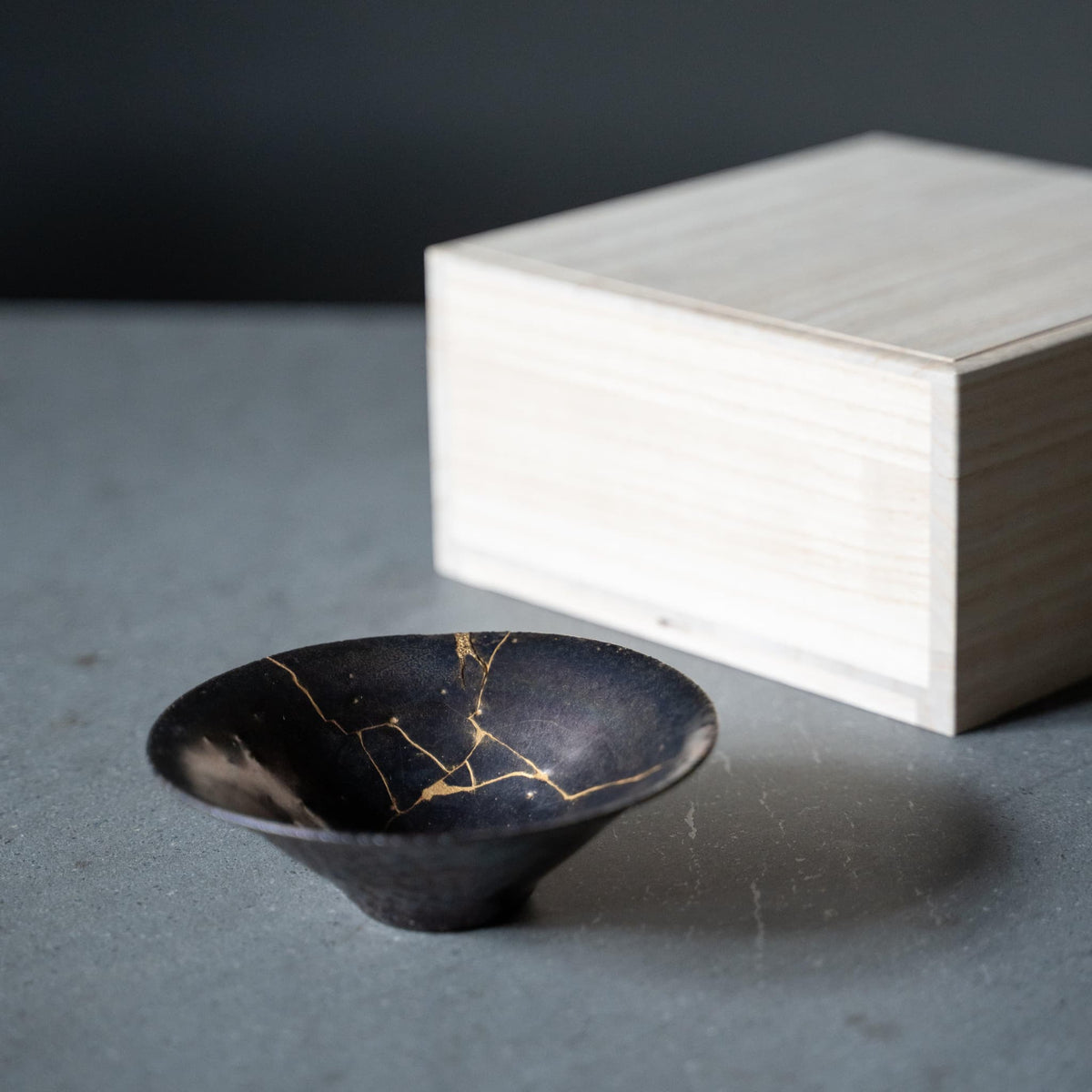 "Japandi-Tischdekoration mit einem grauen Tisch, darauf eine lila Schale, repariert mit Kintsugi, einer traditionellen japanischen Technik, und einer Kiste aus Paulowniaholz."