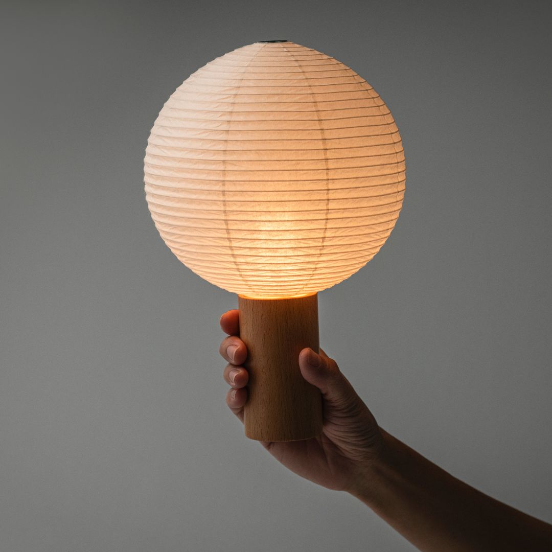 "Tegen een grijze muur staat een draagbare lamp \"TORCHIN\" in het midden aan, gemaakt met de technieken van Yame-chochin lantaarns uit Yame, Fukuoka, Japan, en straalt een warm licht uit. De lampenkap is een ronde lantaarn, en hij heeft een houten cilindrisch handvat dat door een hand wordt vastgehouden."