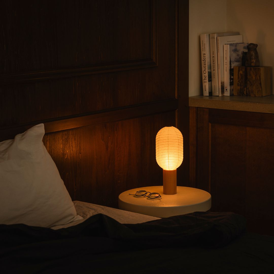 "Une chambre avec un lit à gauche et une étagère avec des livres à droite. Sur la table de chevet à droite du lit se trouve une lampe de chevet en papier « TORCHIN » fabriquée à partir de lanternes japonaises, émettant une lumière chaude de son abat-jour cylindrique."
