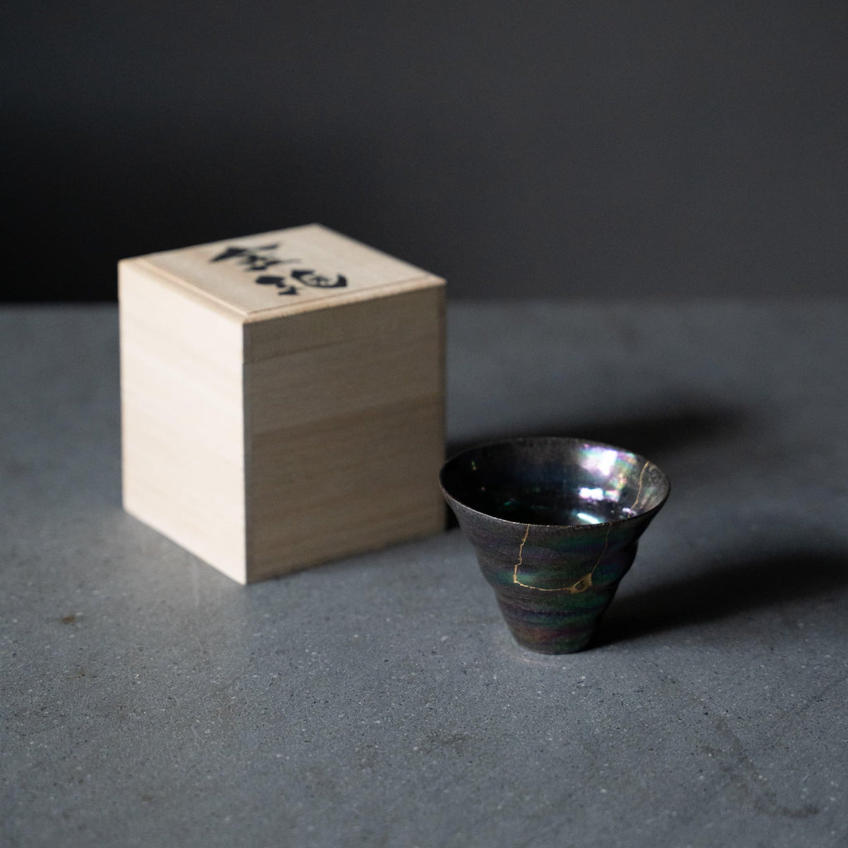 "Kintsugi Sake-Becher „Bunte Ton-Glasur“ und eine Paulownia-Holzschachtel auf grauem Tisch"