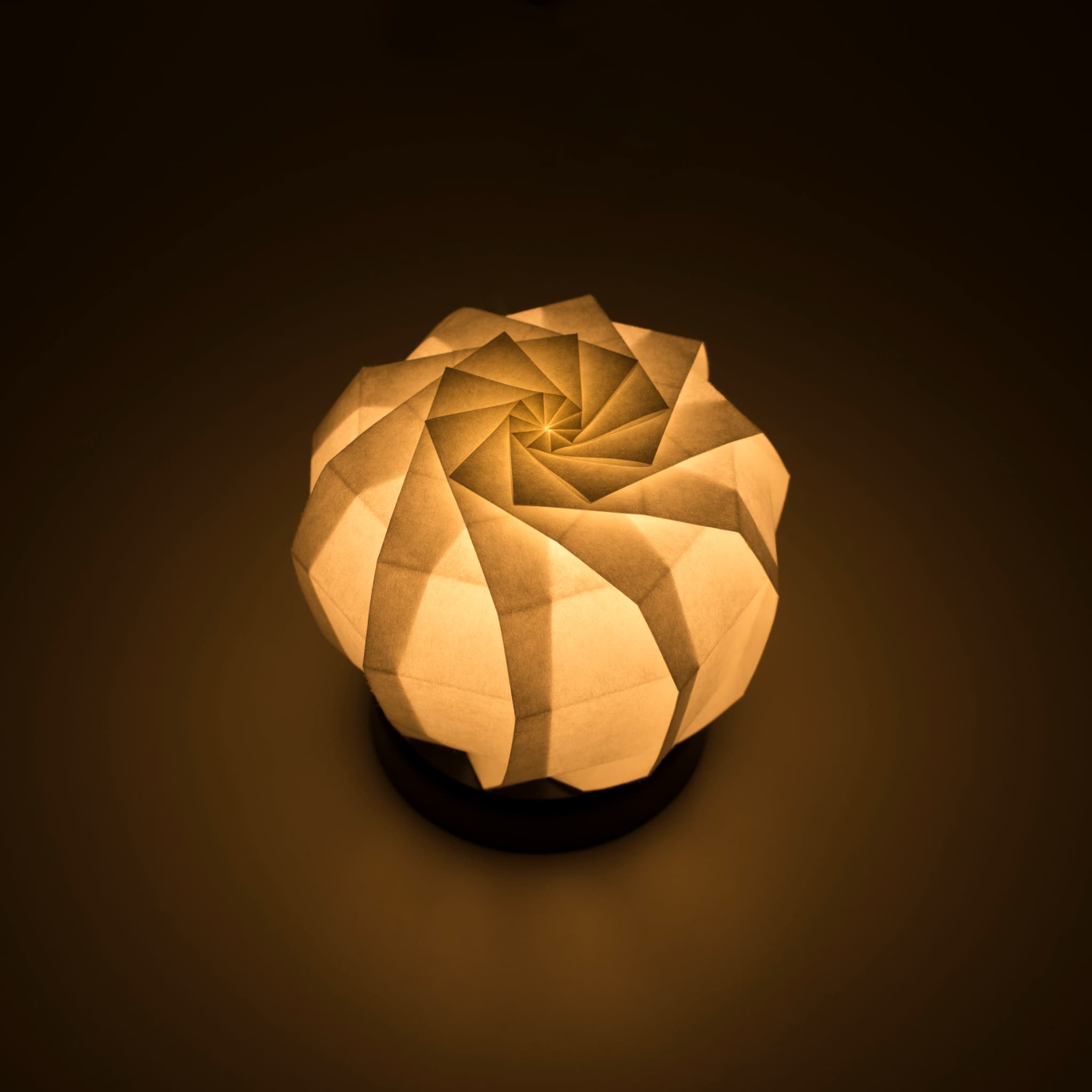 Origami papieren tafellamp met een zwarte voet en een bolvormige witte lampenkap van ingewikkeld gevouwen washipapier, die zacht en warm licht uitstraalt