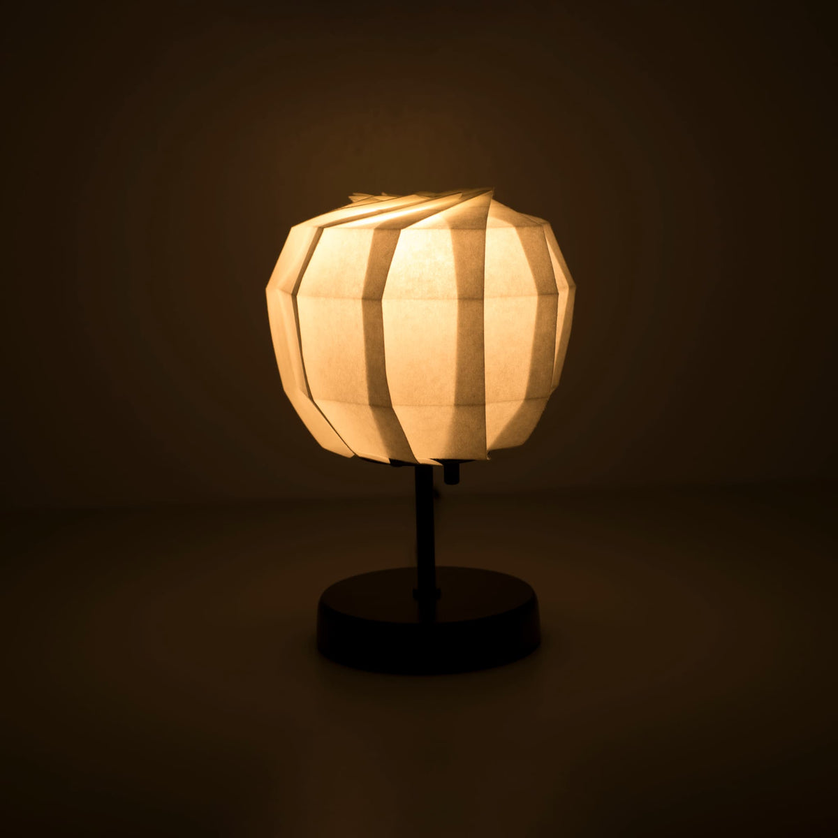 Origami paper table lamp "maru" with a black stand and a spherical white lampshade crafted from intricately folded washi paper, emitting a warm and inviting light.