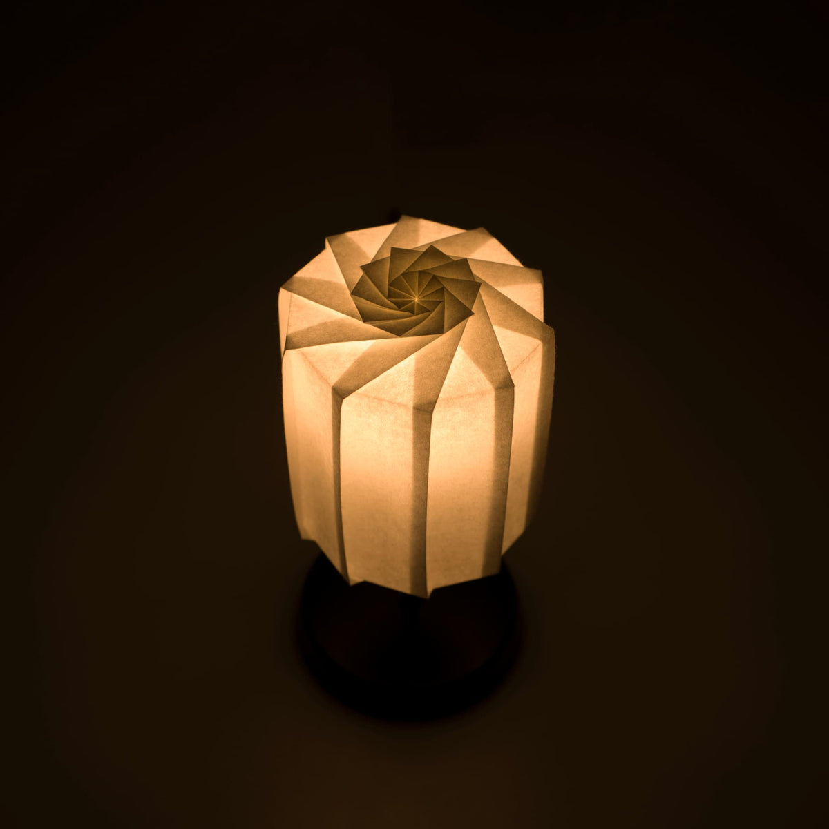 "Lampe de table origami "tutu" avec un pied noir et un abat-jour cylindrique blanc en papier washi plissé, diffusant une lumière chaude et invitante."