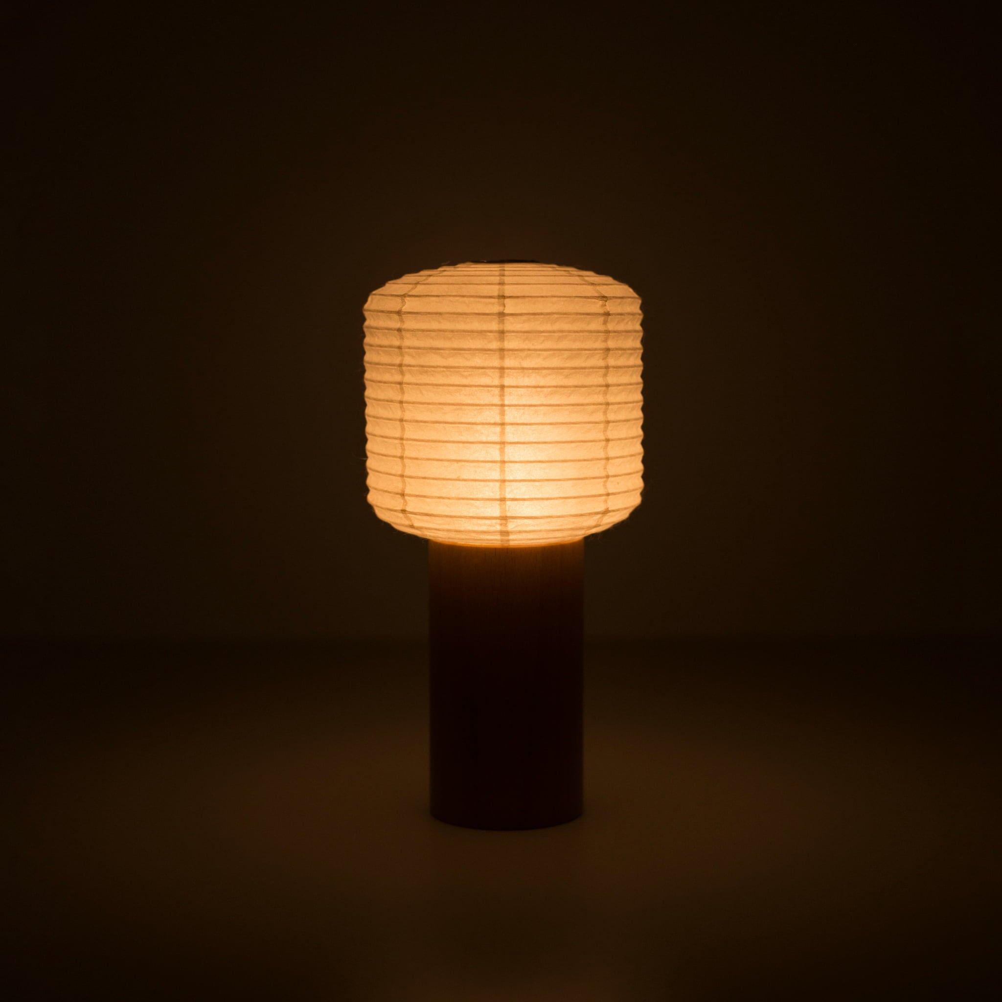 Eine tragbare japanische Papierlampe mit Holzgriff und quadratisch-zylindrischem Papierkopf, die in einem dunklen Raum warmes Licht ausstrahlt