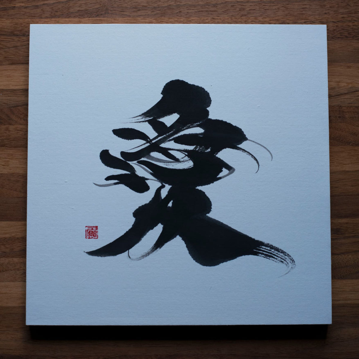 Direct top-down view of a square panel featuring '愛' (love) in Japanese calligraphy, placed on a wooden table.