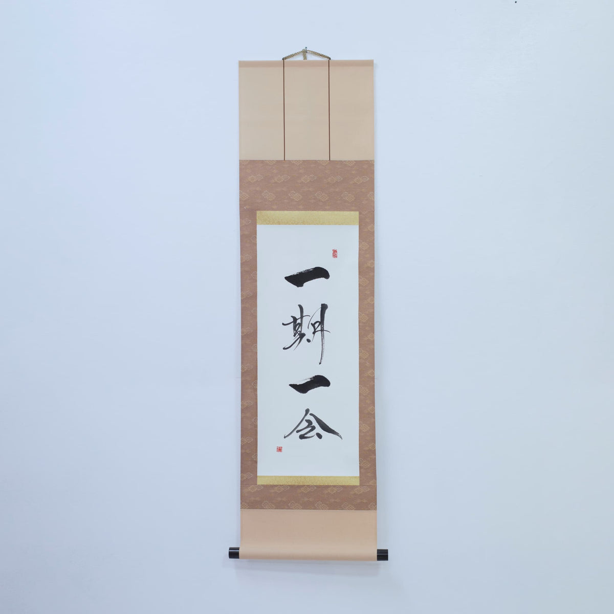 Brown hanging scroll with Japanese calligraphy of '一期一会' (one-time, one meeting) on white rice paper, mounted on a white wall