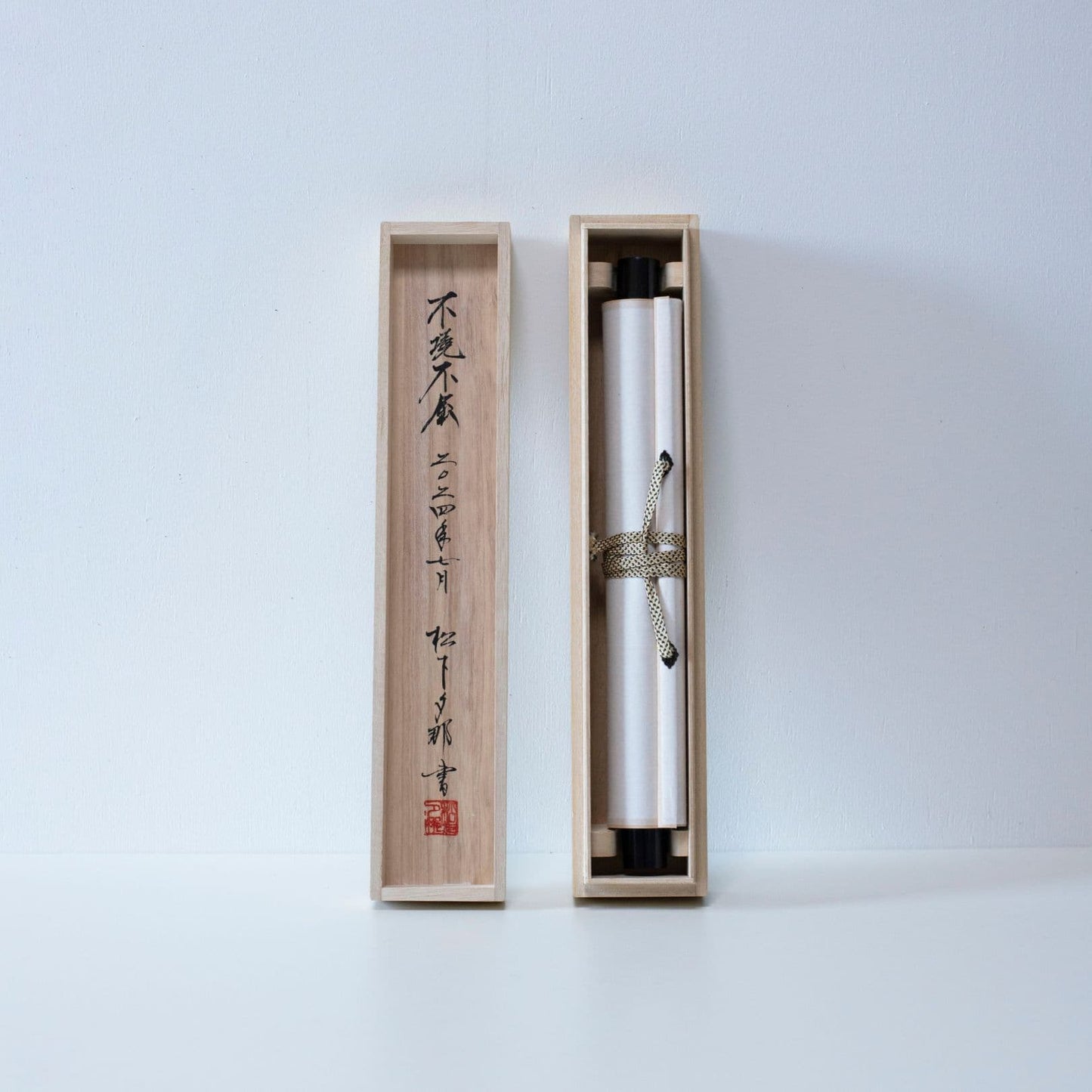 Eine lange, schlanke Holzkiste mit Deckel, die zur Aufbewahrung einer traditionellen japanischen Hänge-Schriftrolle dient und die aufgerollte Schriftrolle im Inneren zeigt