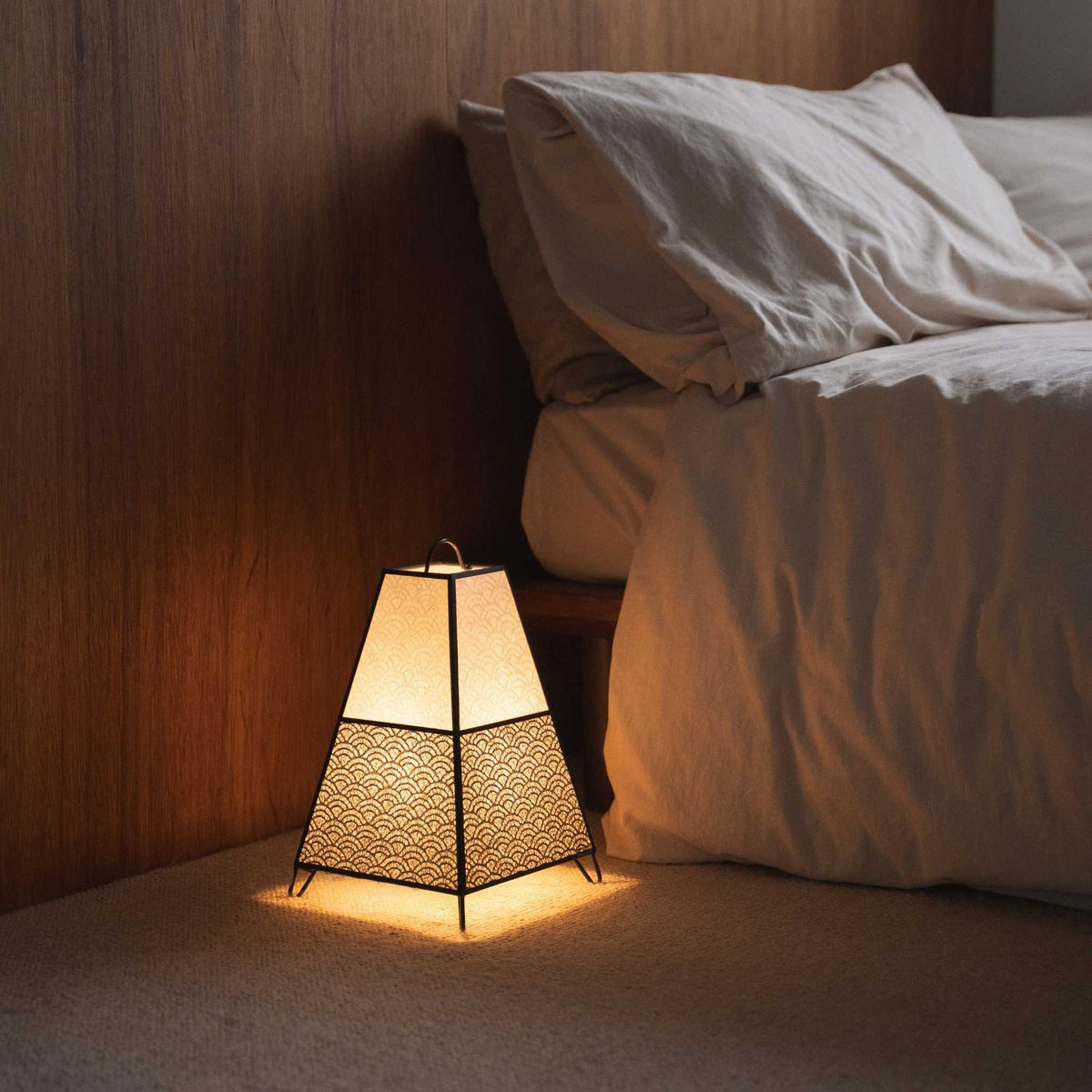 "Lit Japanse vloerlamp KURA Small werpt ingewikkelde schaduwen van zijn papieren kap als sfeerverlichting in een Japandi slaapkamer."