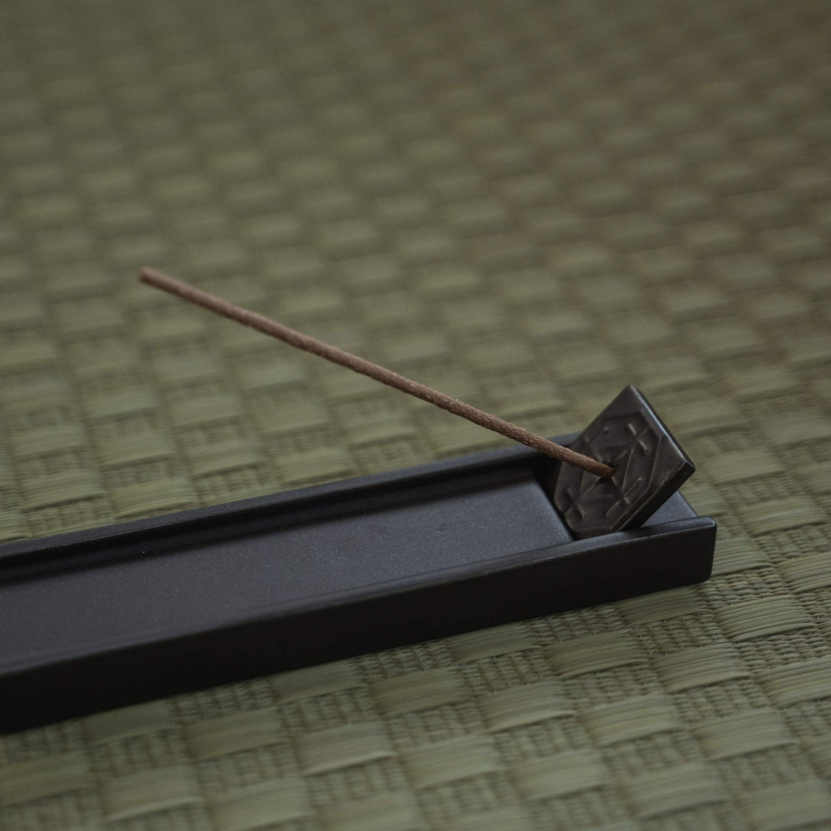 Rectangular dark japanese incense holder on a tatami mat with a burning stick creating a calm atmosphere.