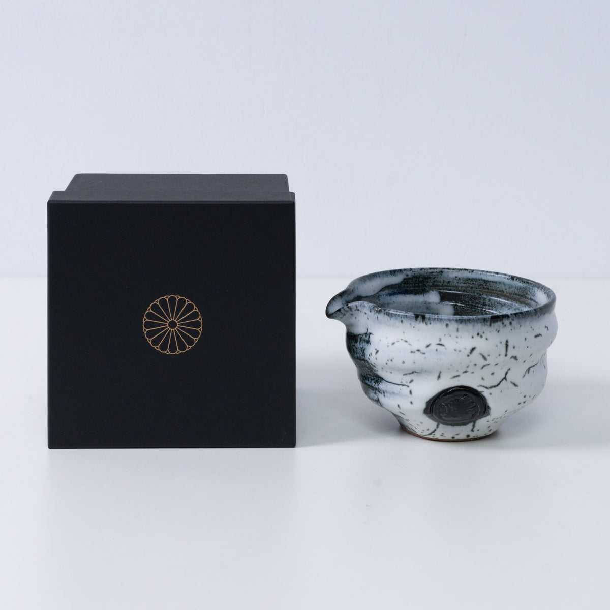 White and black Japanese matcha bowl with spout "Creative Cloud "pottery beside a black gift box with a gold chrysanthemum crest, from Japan.