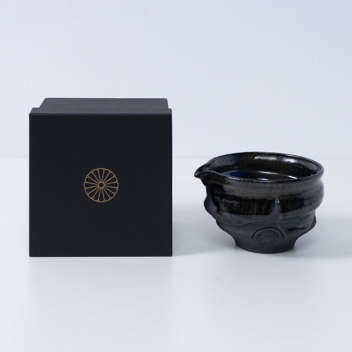 Bol à matcha japonais avec bec verseur "Dignité Bleue" en poterie, à côté d'une boîte cadeau noire avec un blason de chrysanthème doré, du Japon.