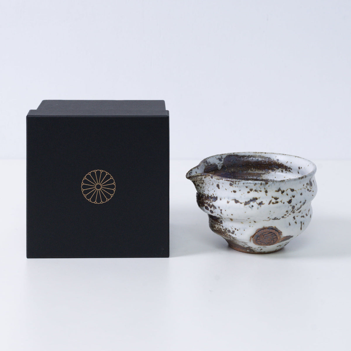 Brown and white Japanese matcha bowl with spout "Zui" pottery next to a black gift box with a gold imperial chrysanthemum crest, from Japan.