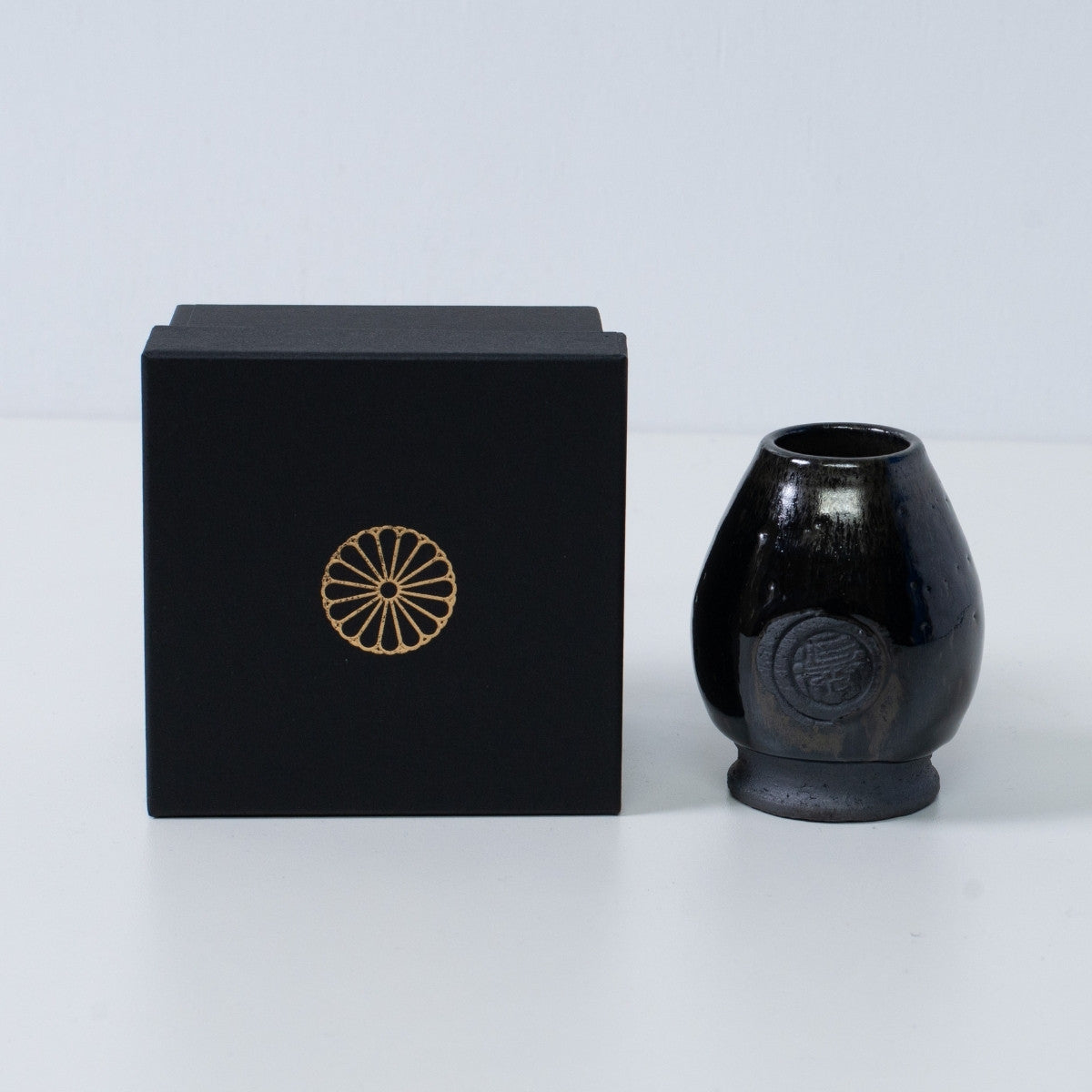 Tiefblauer japanischer Matcha-Schneebesenständer "Dignity Blue" aus Keramik neben einer schwarzen Geschenkbox mit goldenem Chrysanthemenwappen, aus Japan.