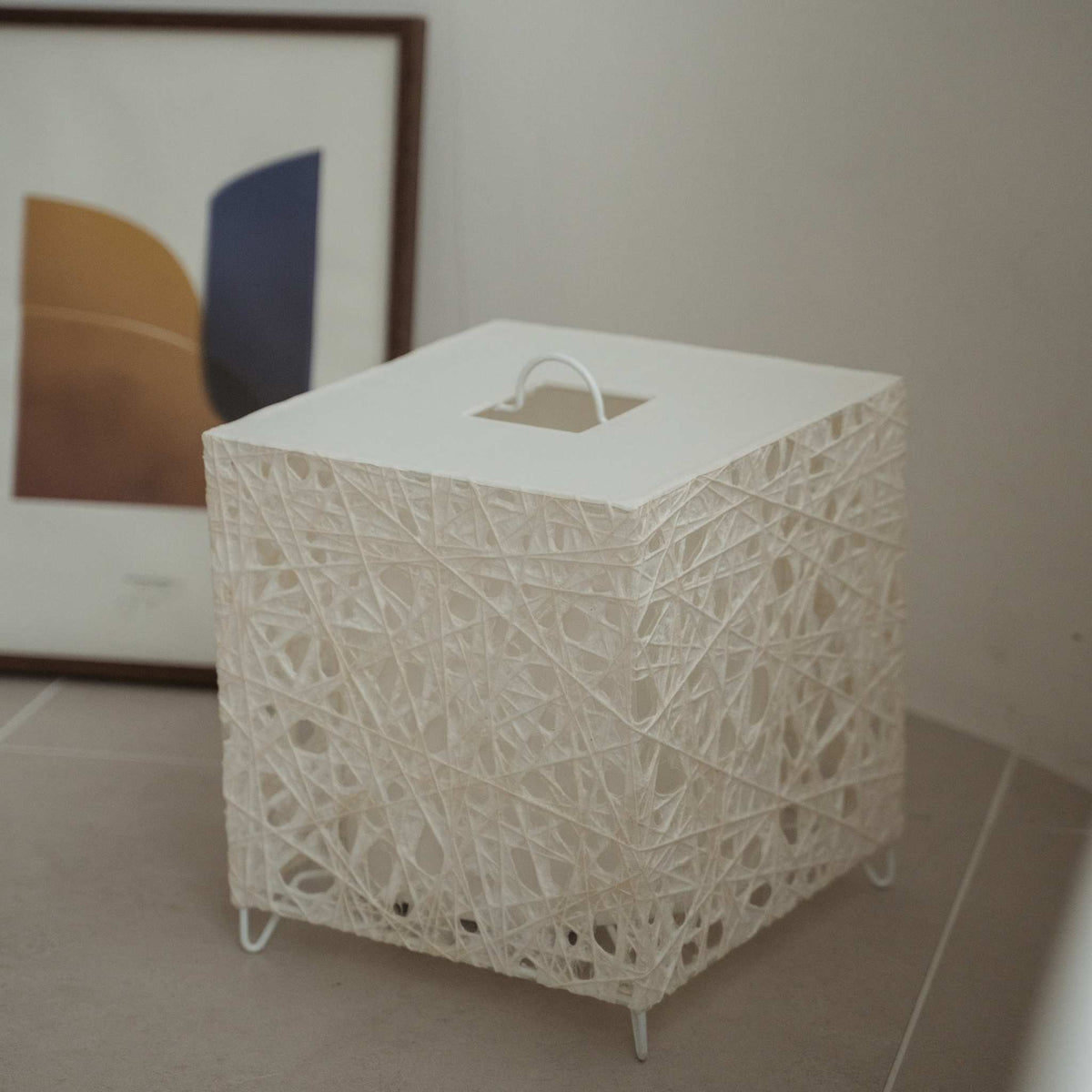 Uoplyst hvid terning japansk gulvlampe Me Look 03 stående på et beige flisegulv ved siden af et indrammet kunsttryk