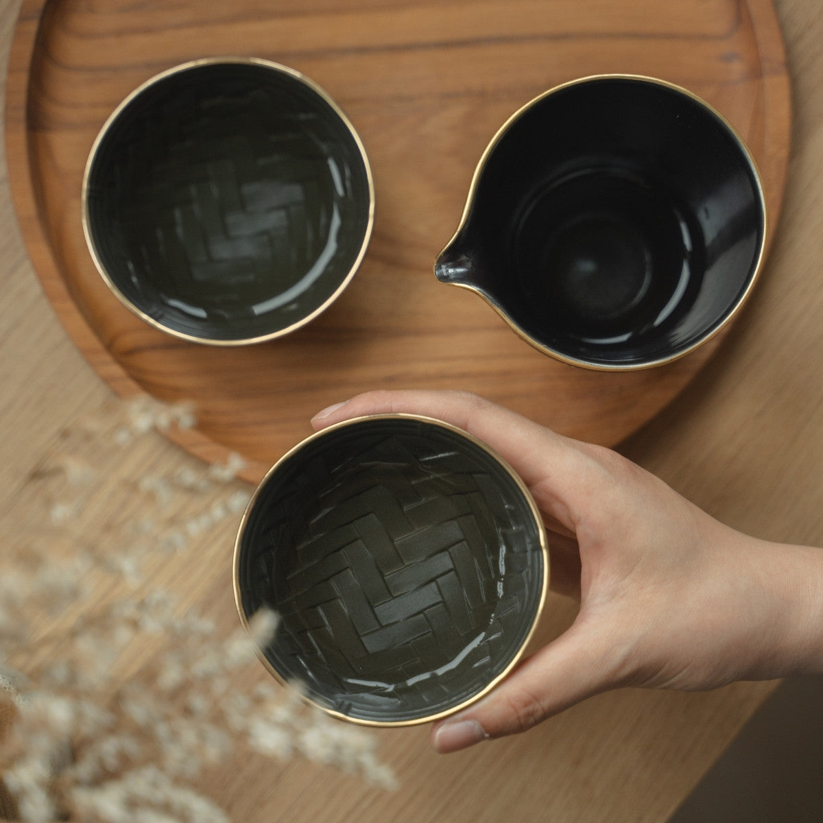 Hand hält eine schwarz-goldene Sake-Schale mit Korbflechtstruktur über einem Tablett, bereit für einen achtsamen Abend.