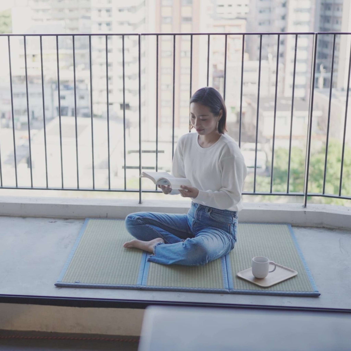 Woman reading a book while sitting on a three panel asanoha tatami mat on a balcony enjoying a zen break.