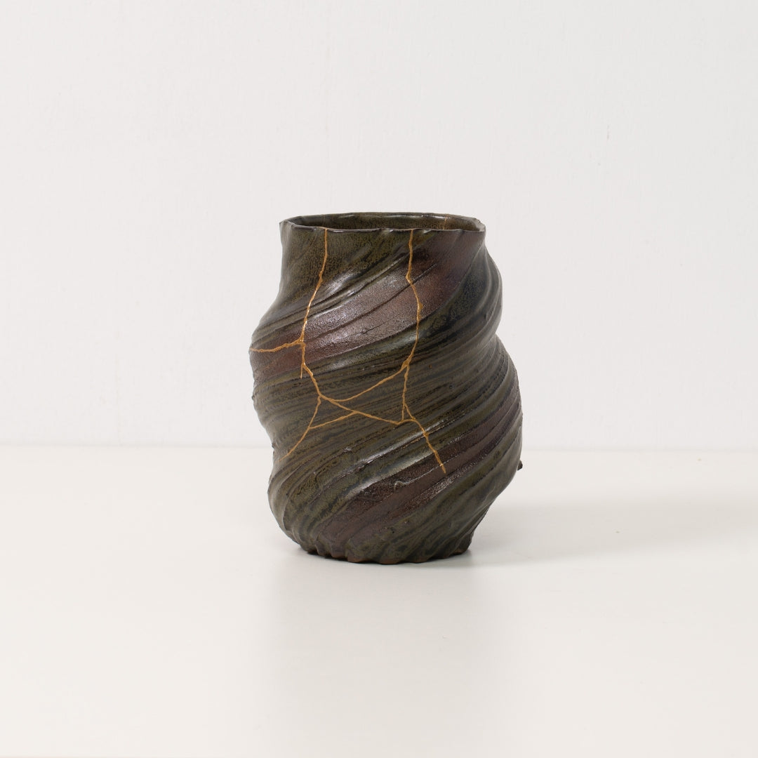Een gedraaide donkere kintsugi-vaas staat alleen op een witte tafel en toont de gouden lijnen van authentieke kintsugi-aardewerkkunst