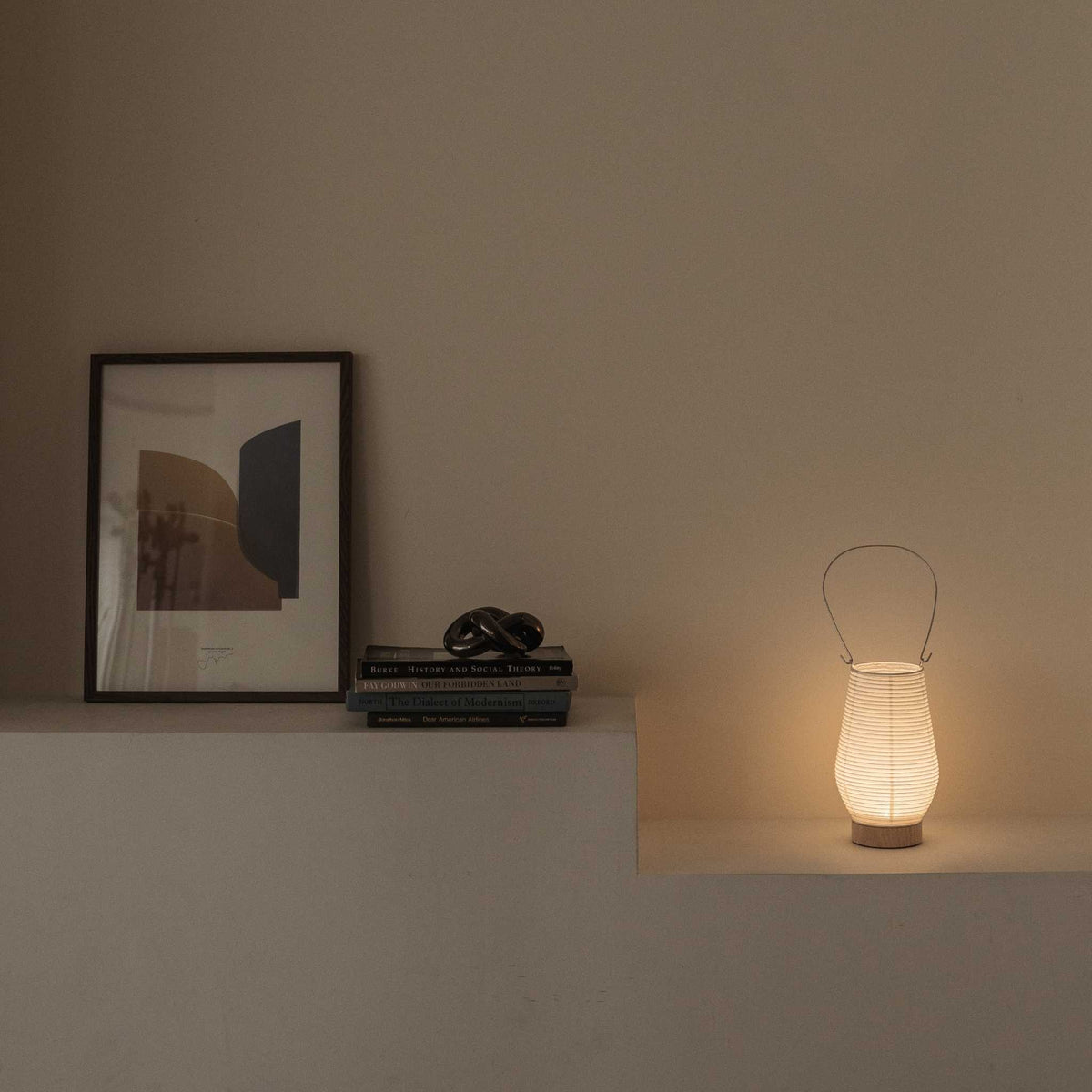 Lysende japansk papir bordlampe RIN 02 på en hylde ved siden af bøger, der giver blødt, stemningsfuldt lys, velegnet til et rum i Japandi-stil