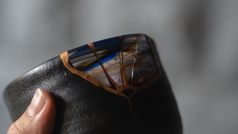 hånd holder en kulsort Kintsugi-tekop Orion med guldfarvede linjer og blåt glas i en wabi-sabi keramikstil.