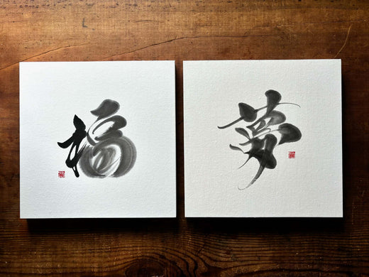 Eine Draufsicht von oben auf zwei quadratische Tafeln mit den japanischen Schriftzeichen für „Glück“ (福) und „Traum“ (夢) in Kalligrafie, nebeneinander auf einem Holztisch liegend.