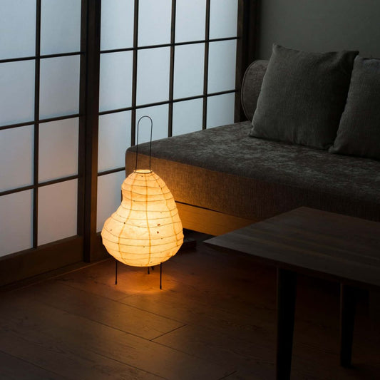 Ein schwach beleuchtetes Zimmer im japanischen Stil mit Shoji-Bildschirmen an der linken Wand und einem grauen Sofa auf der rechten Seite. Eine bohnenförmige japanische Papierbodenlampe, "Beans light", strahlt in der Mitte des Fotos ein warmes Licht aus.