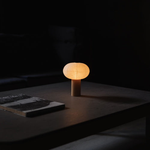 Japanische Papier Stehleuchte "TORCHIN OVAL" mit warmem Licht auf einem grauen Tisch in einem schwach beleuchteten Raum.