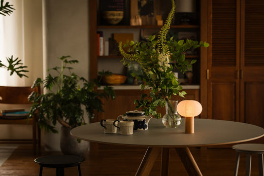 "Ein grauer Tisch mit Holzrand. Auf dem Tisch stehen eine Tischlampe „TORCHIN“ aus japanischen Yame-Chōchin-Laternen, ein Teeservice und eine Glasvase mit einer Zimmerpflanze. Die tragbare Lampe ist angezündet und strahlt ein warmes Licht aus."