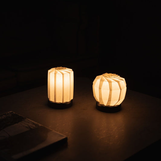 "Zwei Arten von japanischen Origami-Papier-Tischlampen „Blume des Lichts“ strahlen einen warmen Schein und eine ruhige Atmosphäre in einem dunklen Raum aus"