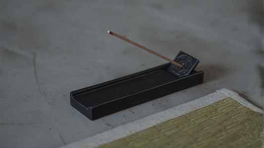 A burning japanese incense stick rests in a black japanese incense burner on a floor next to a traditional tatami mat.