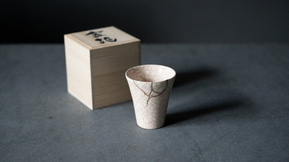 Hvid kintsugi-kop med røde revnemønstre står ved siden af en trækasse, en meningsfuld japansk gave, der repræsenterer wabi sabi.