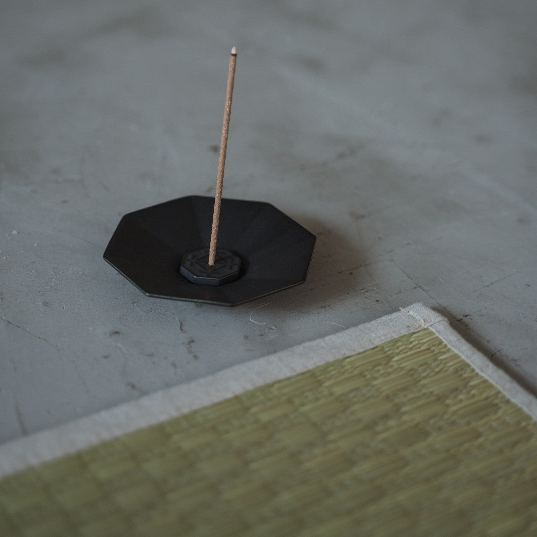 A vertical incense stick stands in an octagon black porcelain burner on a floor beside a Japanese tatami yoga mat.