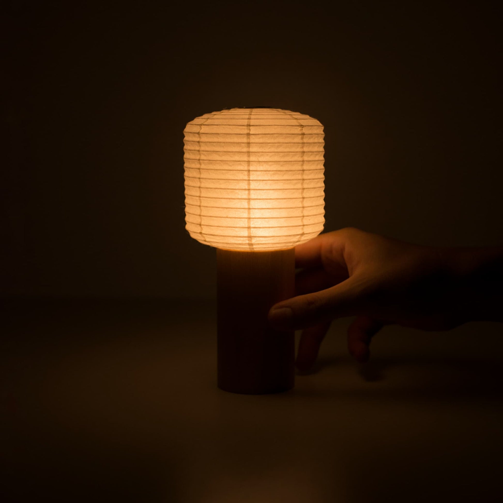 "Eine Hand, die eine warm leuchtende tragbare Papier-Tischlampe in einem dunklen Raum hält"