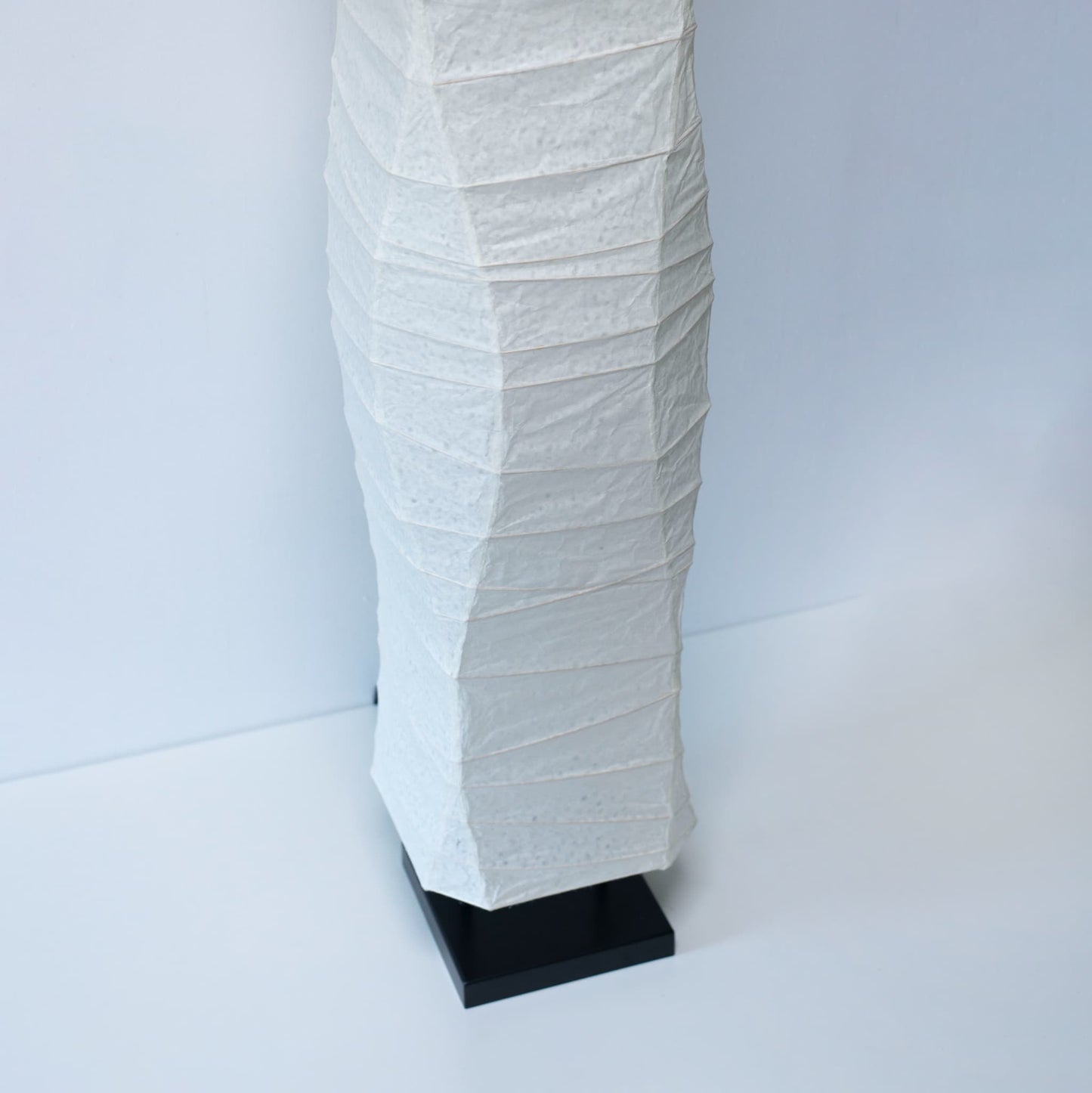 Vue en gros plan, sous un angle, de la partie inférieure d'une lampe en papier japonais washi, montrant son design épuré sur fond blanc
