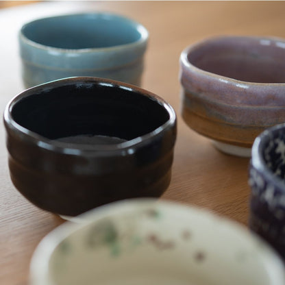 samling af fem håndlavede japanske Mino Ware matcha-skåle i forskellige farver, herunder sort, blå og lilla, placeret på et lyst træbord.
