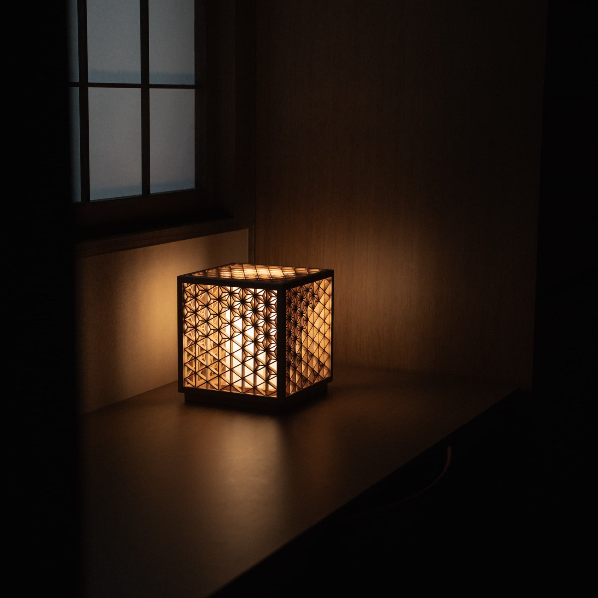 "Lampe Kumiko, méticuleusement fabriquée à la main avec 600 pièces de cèdre Akita de première qualité, diffusant une lumière chaude et invitante sur une table de chevet dans une chambre japonaise traditionnelle."