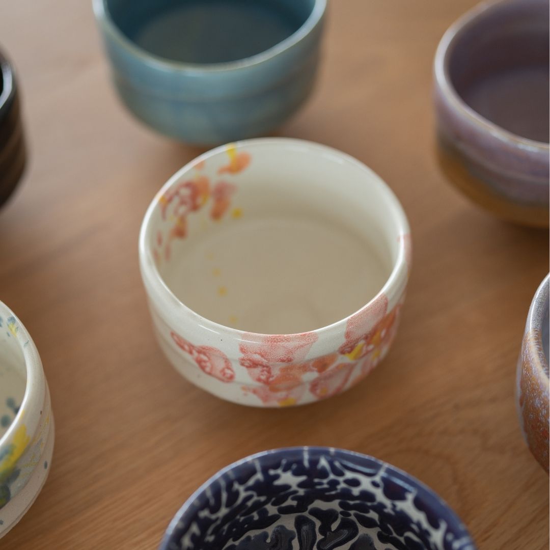 Et nærbillede af en hvid Mino Ware matcha chawan-skål med delikat lyserød og orange blomsterglasur, omgivet af andre farverige håndlavede japanske matcha-skåle på en træoverflade.