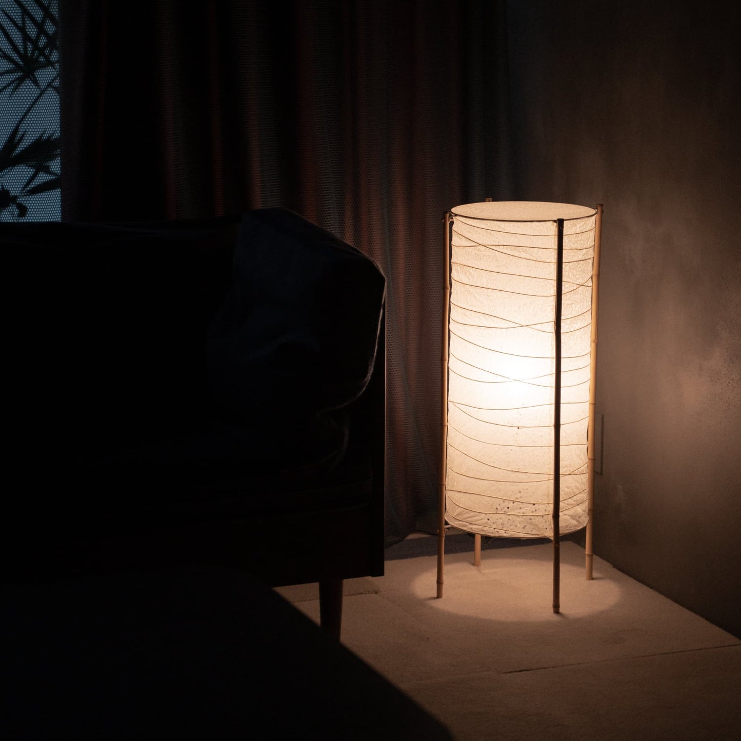 Lampe sur pied cylindrique en papier japonais washi avec cadre en bambou, placée dans le coin d'une pièce faiblement éclairée, illuminant doucement un canapé voisin dans un salon Japandi.