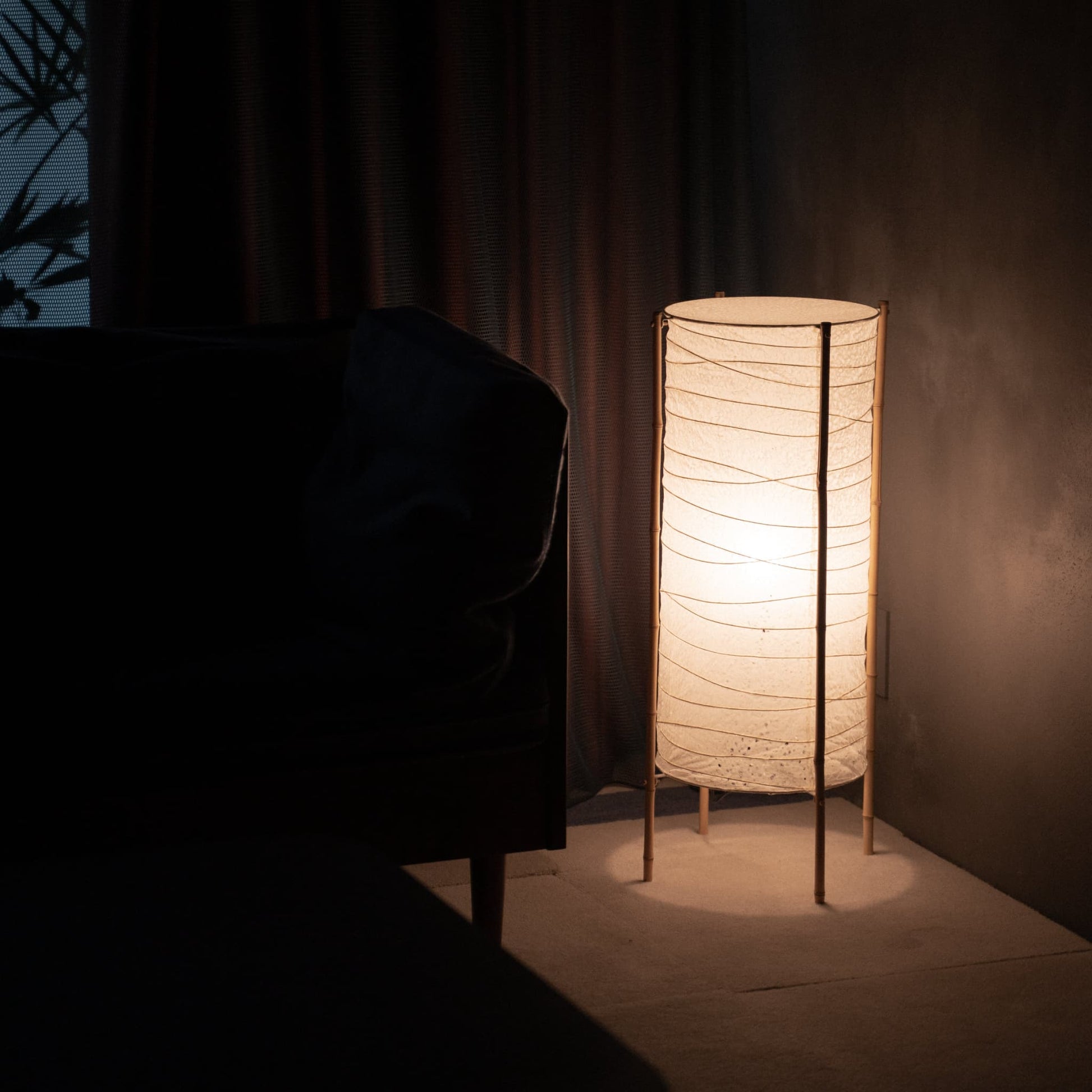 Lampe sur pied cylindrique en papier japonais washi avec cadre en bambou, placée dans le coin d'une pièce faiblement éclairée, illuminant doucement un canapé voisin dans un salon Japandi.