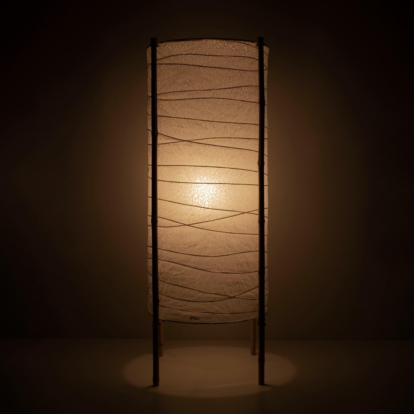 "Vue complète d'une lampe japonaise cylindrique avec abat-jour en papier washi et cadre en bambou, lumière chaude sur fond marron"