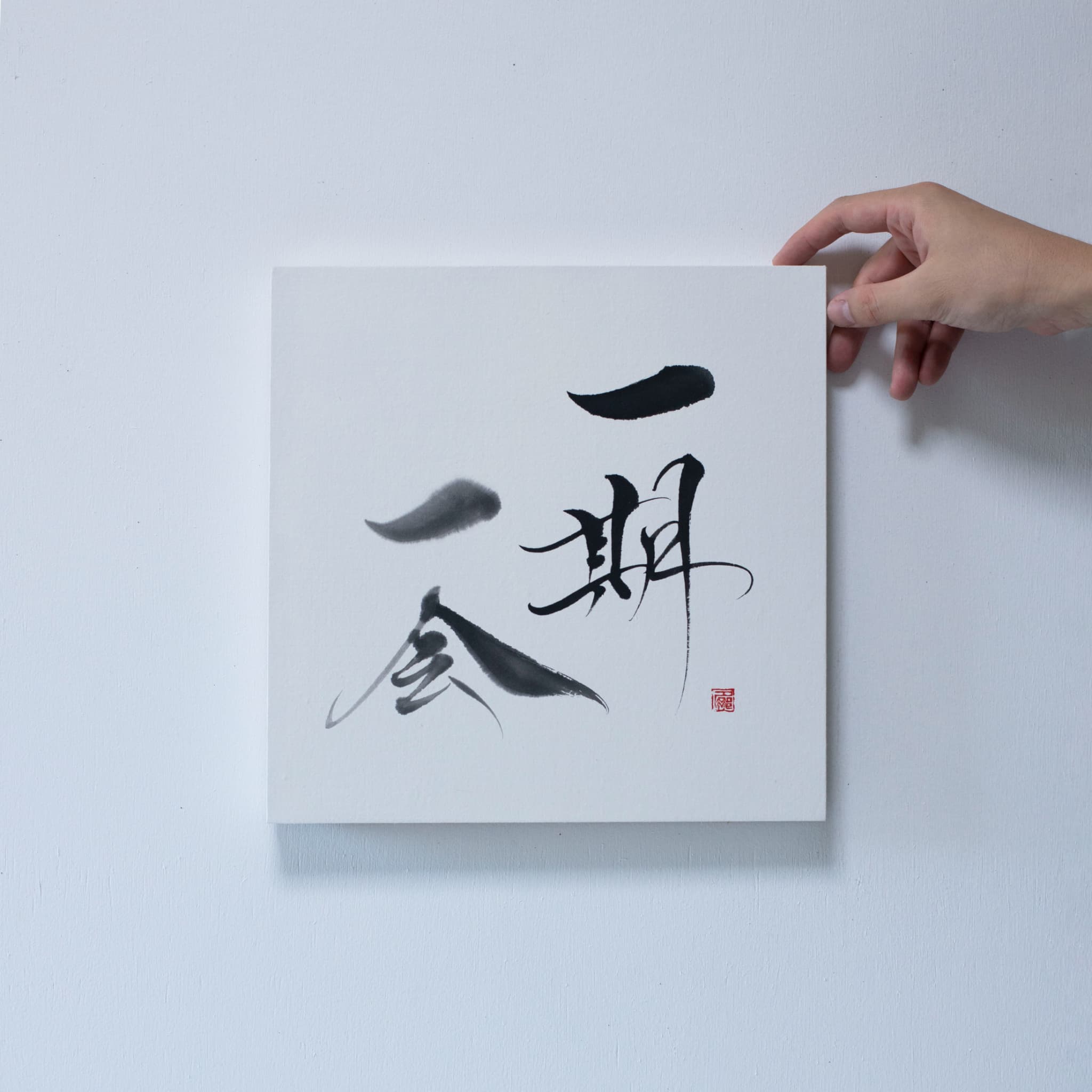 Gros plan d'une main tenant le coin d'un panneau carré affichant « Ichigo Ichie » en calligraphie japonaise, fixé sur un mur blanc