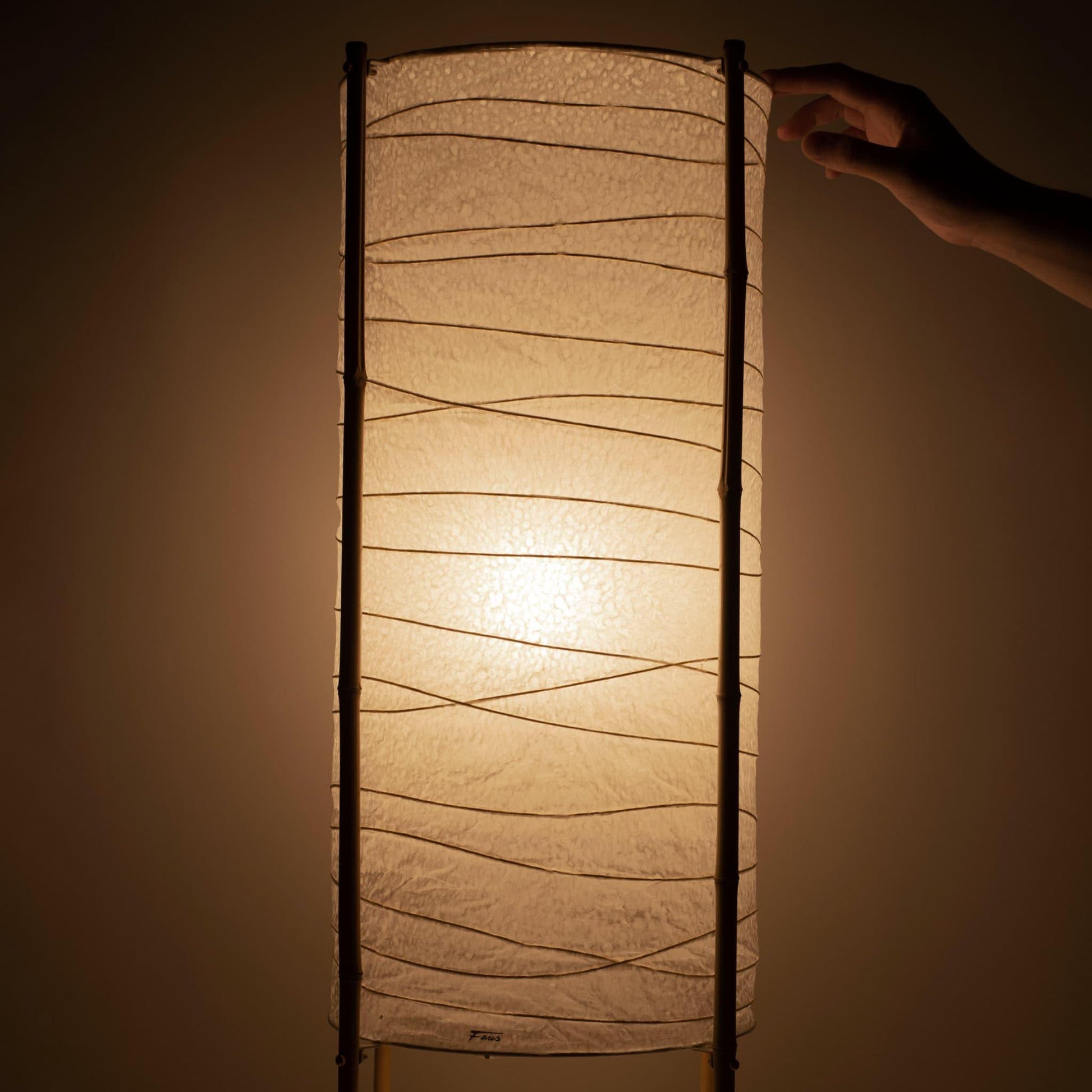 Gros plan d'une main touchant le dessus d'une lampe cylindrique japonaise en papier washi avec un cadre en bambou, lumière chaude sur fond marron