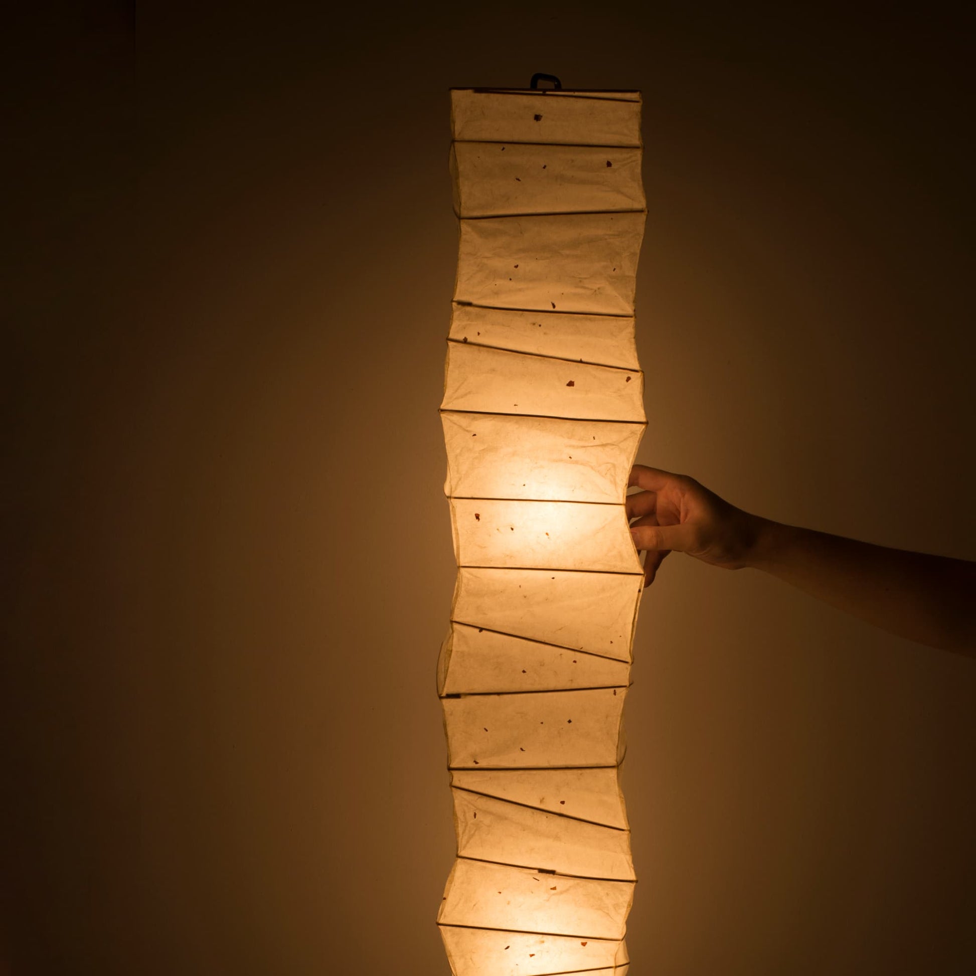Gros plan d'une main touchant le côté d'une lampe en papier japonais washi incurvé, lumière chaude sur fond marron