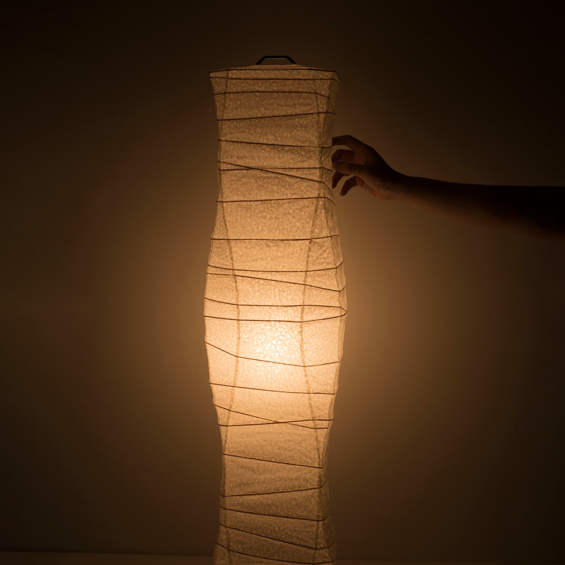 "Une main touche délicatement le côté d'une lampe en papier japonais washi aux courbes fluides, émettant une douce lumière chaude"