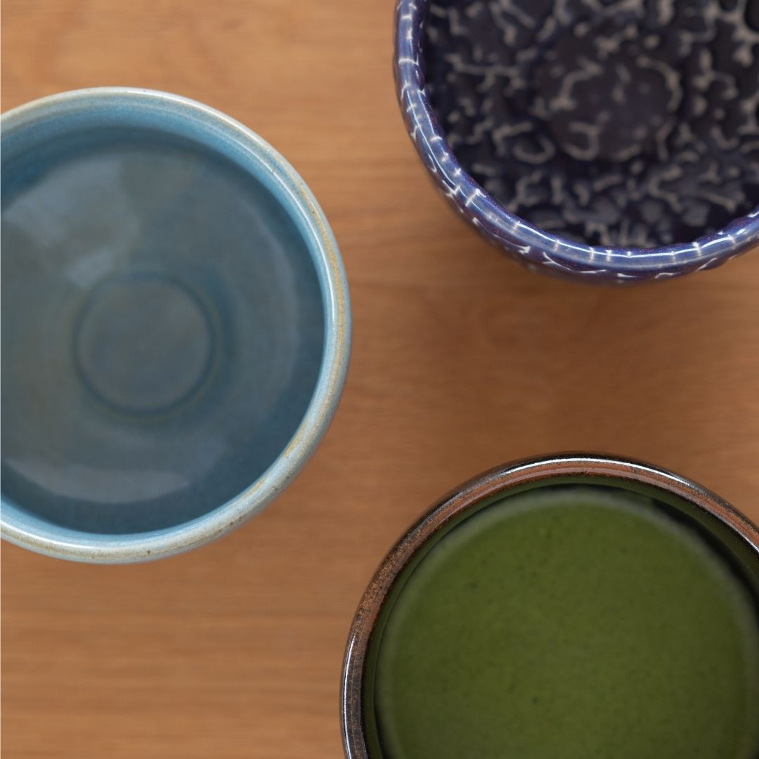 Overheadbillede af tre håndlavede japanske matcha chawan-skåle (blå, mørkeblå Kairagi og sort) på et træbord.