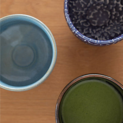 Overheadbillede af tre håndlavede japanske matcha chawan-skåle (blå, mørkeblå Kairagi og sort) på et træbord.
