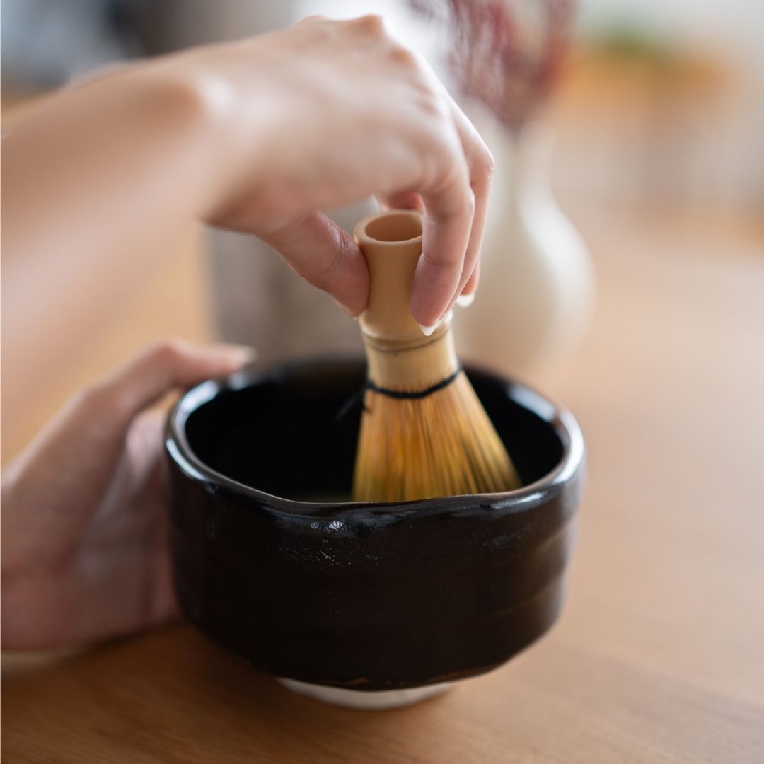 hånd, der holder en bambuspisk, og som tilbereder matcha i en sort håndlavet japansk Mino-skål.