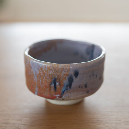 "close-up af en enkelt håndlavet japansk matcha-skål (chawan) med en dristig navyblå og rød splash-glasur over en kinyo lilla base, stående på et træbord."