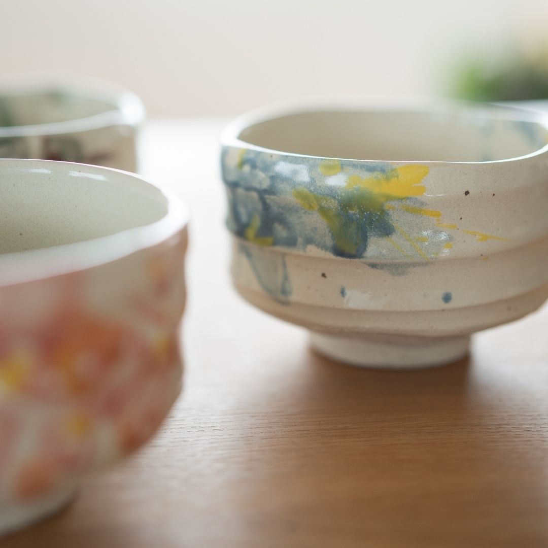 Fokus auf die japanische Matcha-Schale "Splash" mit blauer und gelber Glasur, die auf einem Holztisch neben der rosa Schale "Flower" steht und die Vielfalt handgefertigter Shino-Keramik zeigt.